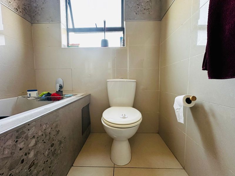 A functional bathroom featuring a toilet, bathtub, and neutral wall and floor tiling with a top-hung window for ventilation.