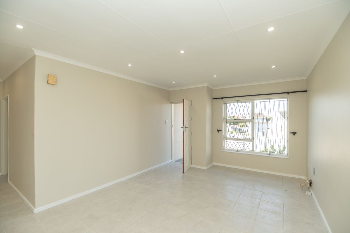 A bright, empty living room featuring neutral walls, tiled flooring, and recessed downlighting, with security bars on the window.