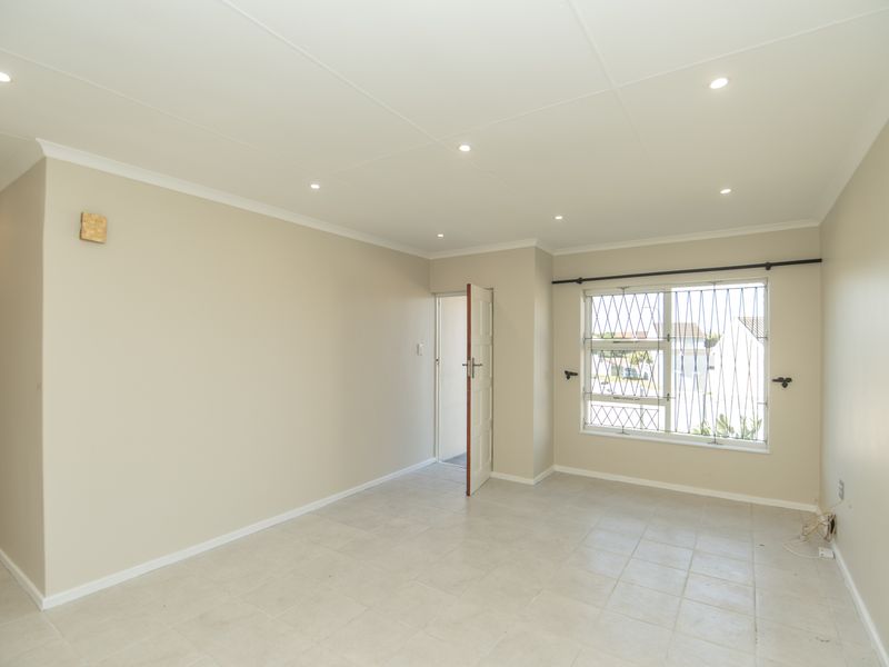A bright, empty living room featuring neutral walls, tiled flooring, and recessed downlighting, with security bars on the window.