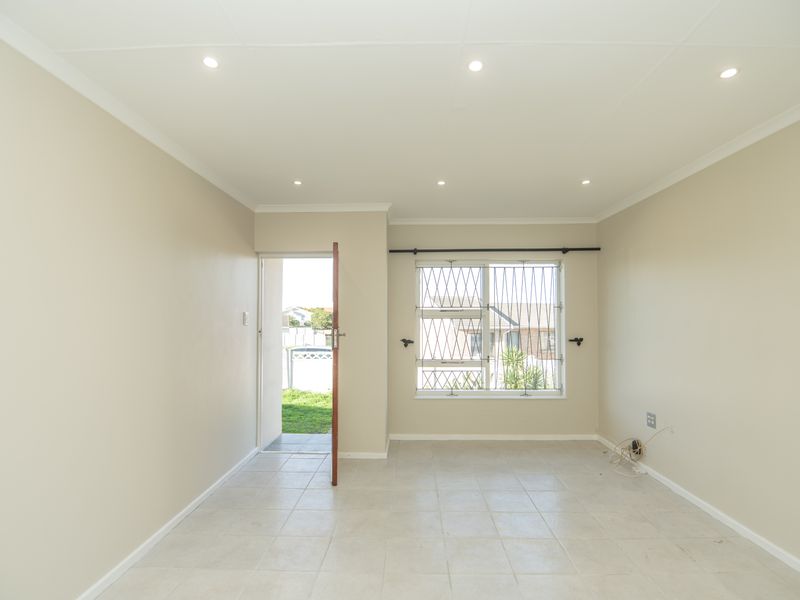 A bright, empty living room featuring neutral walls, tiled flooring, recessed downlighting, and direct access to the rear garden through a security door.