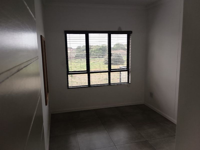 A clean, empty bedroom featuring neutral tiled flooring and a large multi-pane window with fitted venetian blinds, offering a view of the surrounding area.