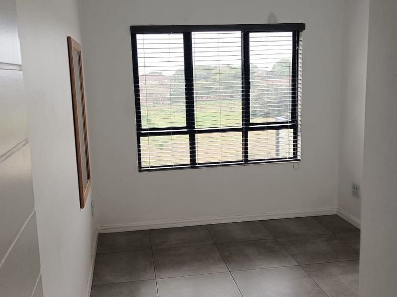 A clean, empty bedroom featuring neutral grey floor tiles, a large window with fitted blinds, and a modern, minimalist aesthetic.