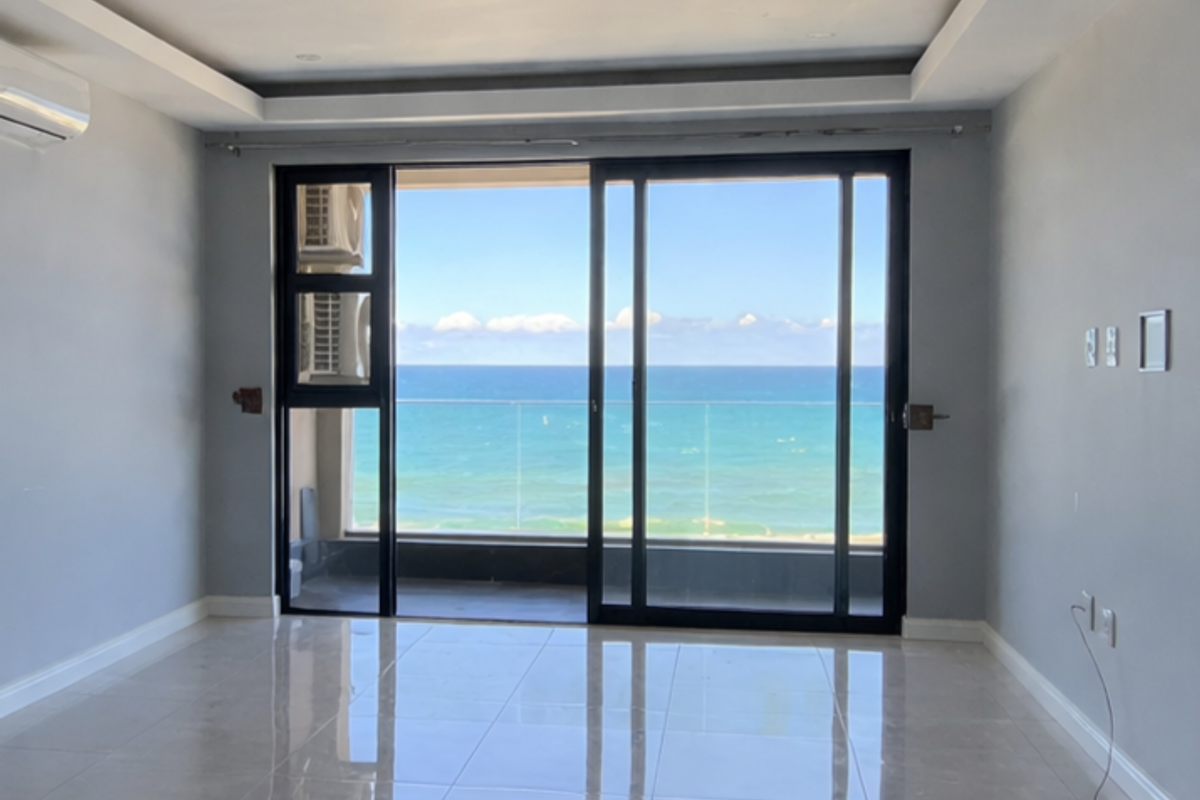 Bright, empty living area featuring polished porcelain tiles, recessed ceiling lighting, and large sliding doors opening to a balcony with expansive ocean views.