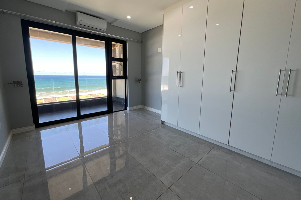 Modern, empty bedroom featuring high-gloss built-in cupboards, large format floor tiles, and a sliding glass door opening to a balcony with ocean views.