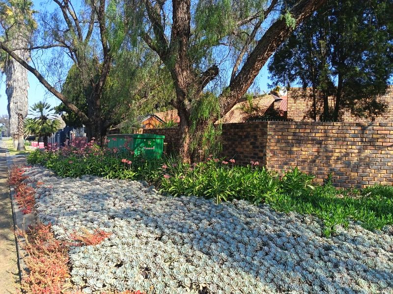 A view of the property's street-facing boundary, featuring a sturdy brick wall with security spikes and a well-maintained landscaped verge with mature trees.