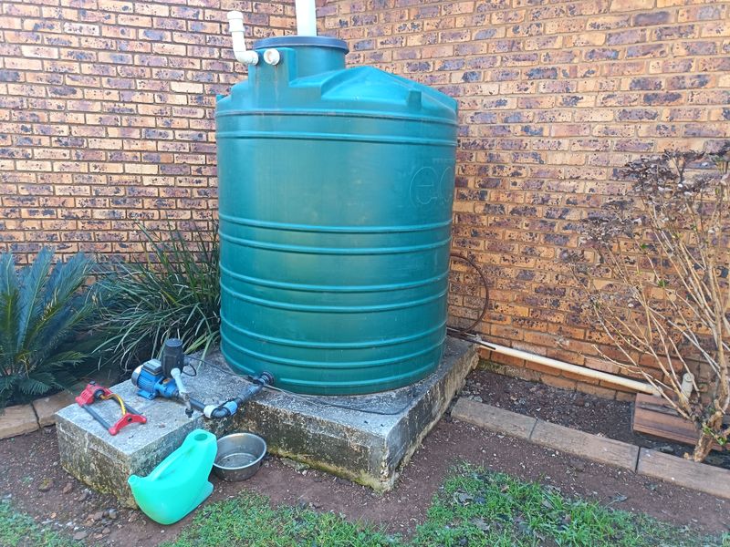 Exterior rear area featuring a green water storage tank mounted on a concrete plinth with an integrated water pump system against a brick wall.