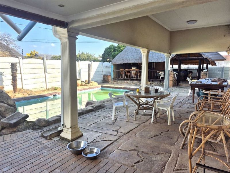 Covered outdoor patio area featuring paved flooring and decorative columns, overlooking a swimming pool and a separate thatched lapa structure.