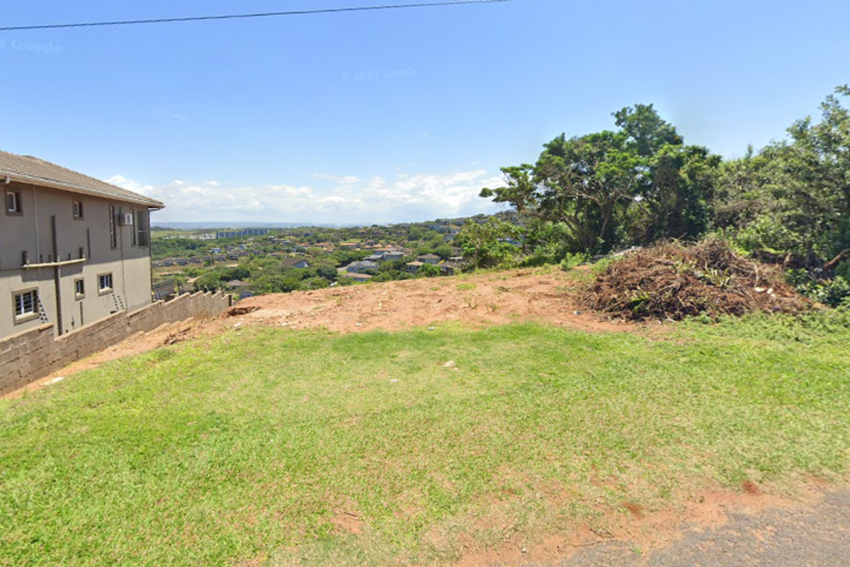 A view of a vacant residential plot featuring sloped terrain and a retaining wall, overlooking the surrounding neighborhood landscape.