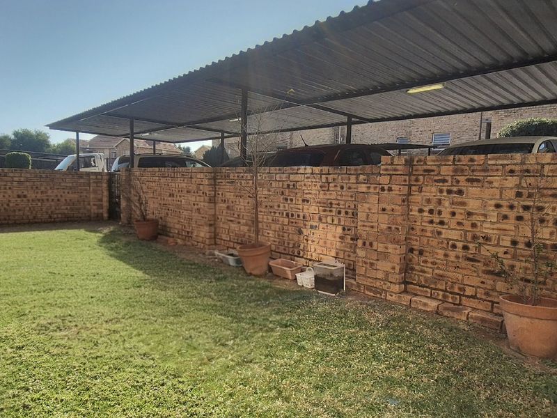 A rear garden area featuring a well-maintained lawn, a sturdy brick boundary wall, and a covered carport structure.