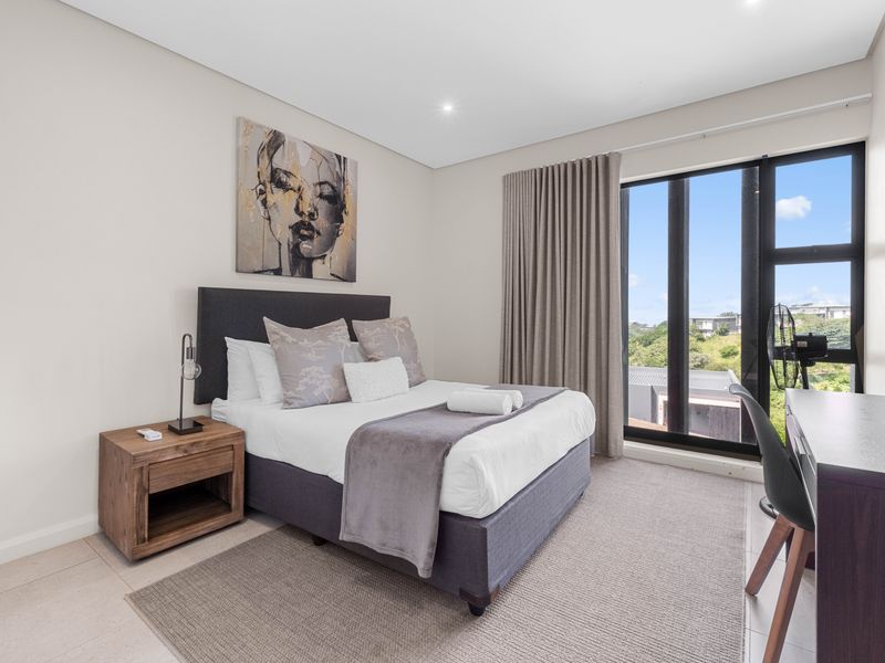 A modern, well-maintained bedroom featuring neutral tones, tiled flooring, recessed lighting, and a large window providing natural light and exterior views.