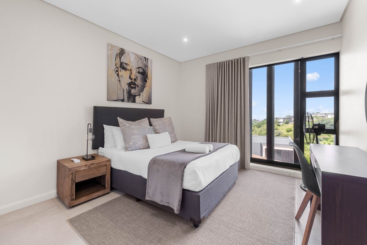 A modern, well-maintained bedroom featuring neutral tones, tiled flooring, recessed lighting, and a large window providing natural light and exterior views.