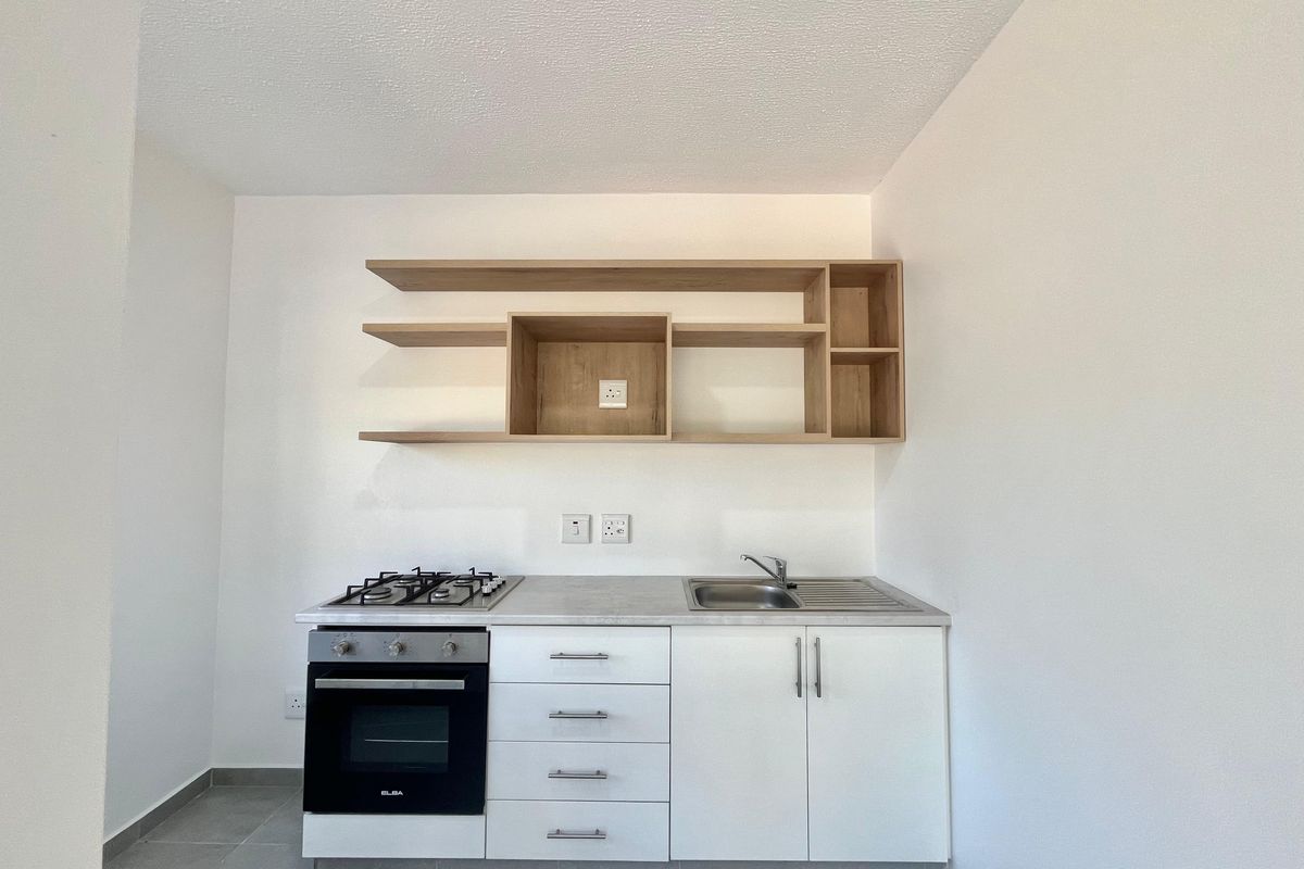 A compact, modern kitchen featuring white base cabinetry, a gas hob, built-in oven, stainless steel sink, and open wooden shelving.
