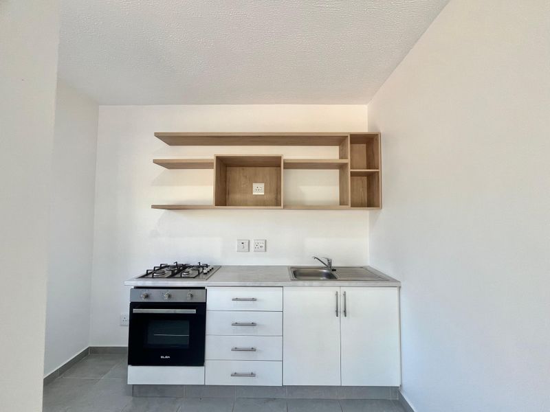 A compact, modern kitchen featuring white base cabinetry, a gas hob, built-in oven, stainless steel sink, and open wooden shelving.
