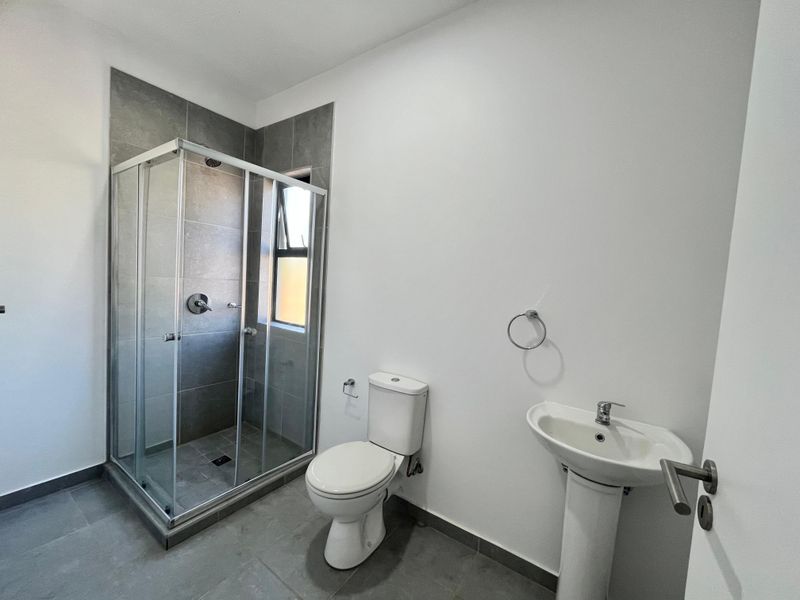 A clean, modern bathroom featuring a glass-enclosed shower, pedestal basin, and toilet with neutral grey floor and wall tiling.