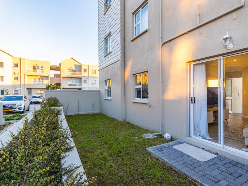 A rear exterior view of the apartment featuring a small private lawn, paved patio area, and sliding glass door access to the living space.