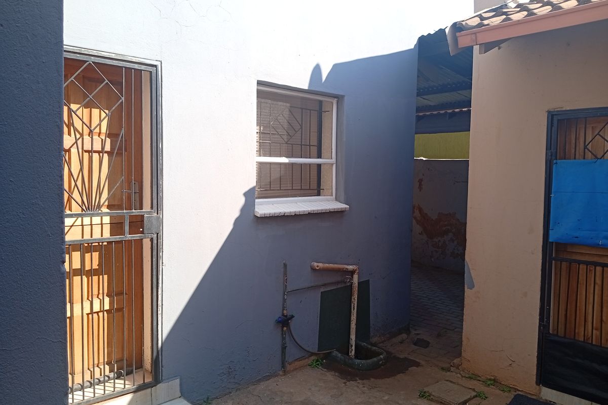 A view of the rear exterior area showing a security gate, window with burglar bars, and exposed external plumbing against a painted wall.