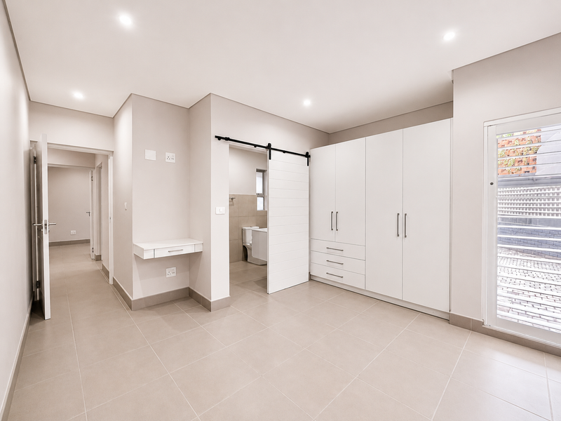 A bright, modern bedroom featuring neutral tiled floors, extensive built-in cabinetry, a sliding barn door leading to an en-suite, and a wall-mounted desk.
