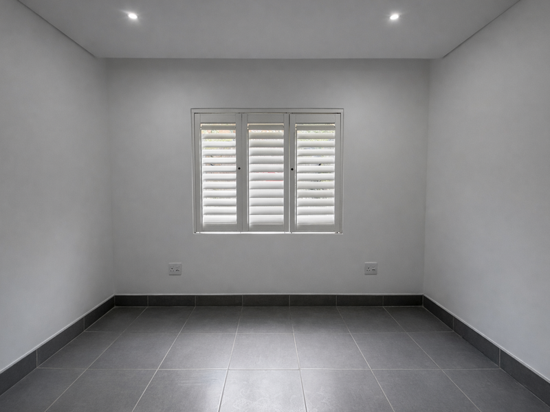 A clean, empty bedroom featuring neutral grey floor tiles, white walls, recessed downlights, and white plantation shutters on the window.