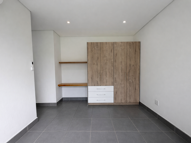 A clean, modern bedroom featuring neutral tiled flooring, recessed lighting, and a functional built-in wardrobe with integrated shelving.