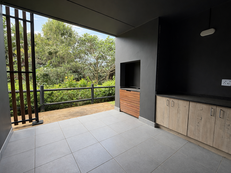 A modern covered patio featuring a built-in braai, timber decking, and custom cabinetry with a stone countertop, overlooking lush greenery.