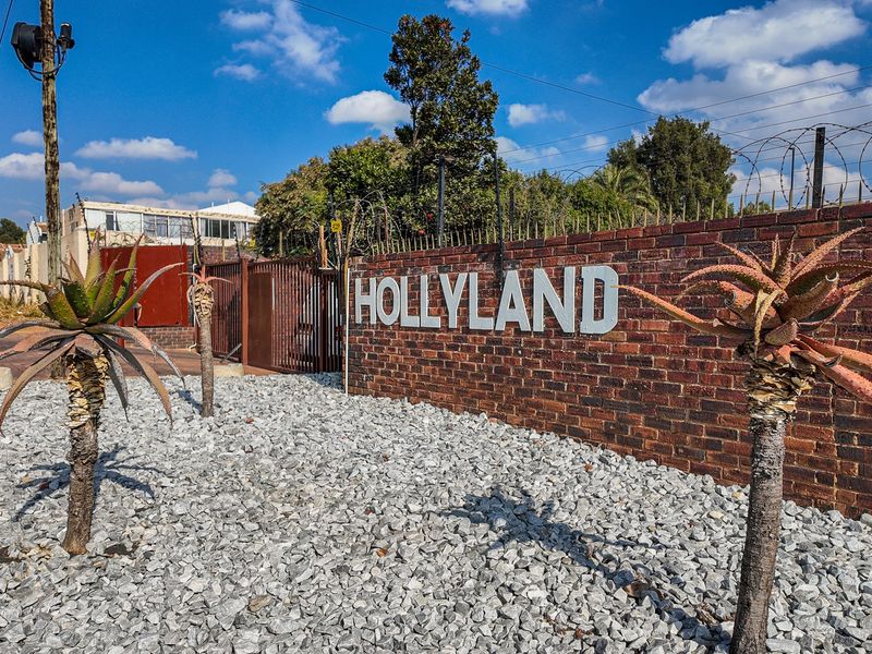 The entrance to the Hollyland complex featuring a brick boundary wall, security gate, and gravel landscaping.