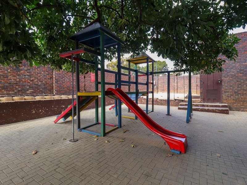 A paved rear courtyard area featuring a brick boundary wall and a fixed children's playground structure shaded by a large tree.