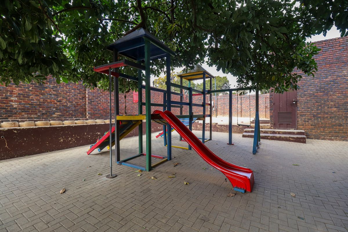 A paved rear courtyard area featuring a brick boundary wall and a fixed children's playground structure shaded by a large tree.