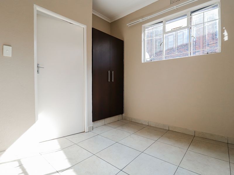 A bright, empty bedroom featuring neutral wall paint, tiled flooring, and a built-in dark wood cupboard.
