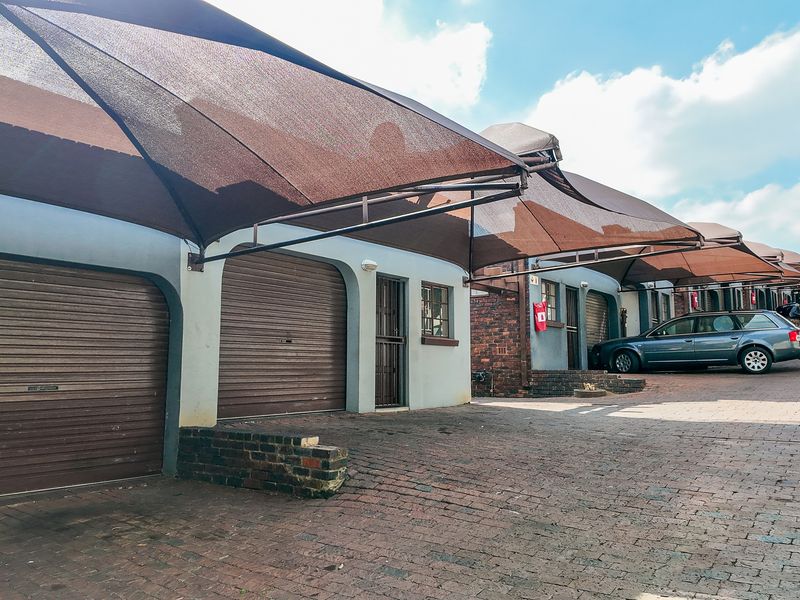 A row of townhouse garages featuring roller shutter doors and a shared overhead shade cloth carport structure on a brick-paved driveway.