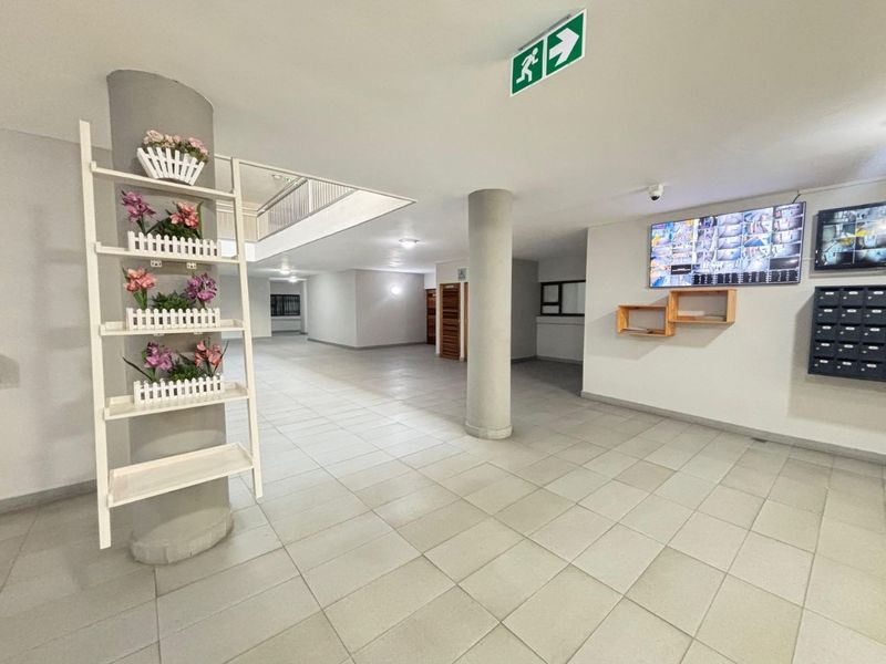 A spacious, well-maintained apartment building lobby or hallway featuring tiled floors, structural columns, security monitoring systems, and integrated mailboxes.