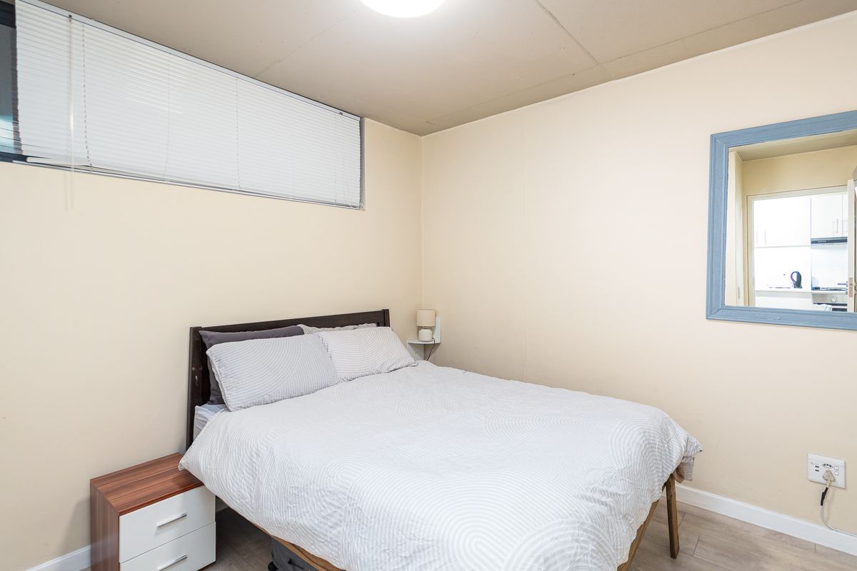 A functional bedroom featuring laminate flooring, neutral wall finishes, and a high-set window with fitted venetian blinds.