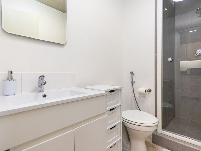 A clean, modern bathroom featuring a white vanity with basin, a wall-mounted mirror, a toilet with bidet spray, and a glass-enclosed shower.