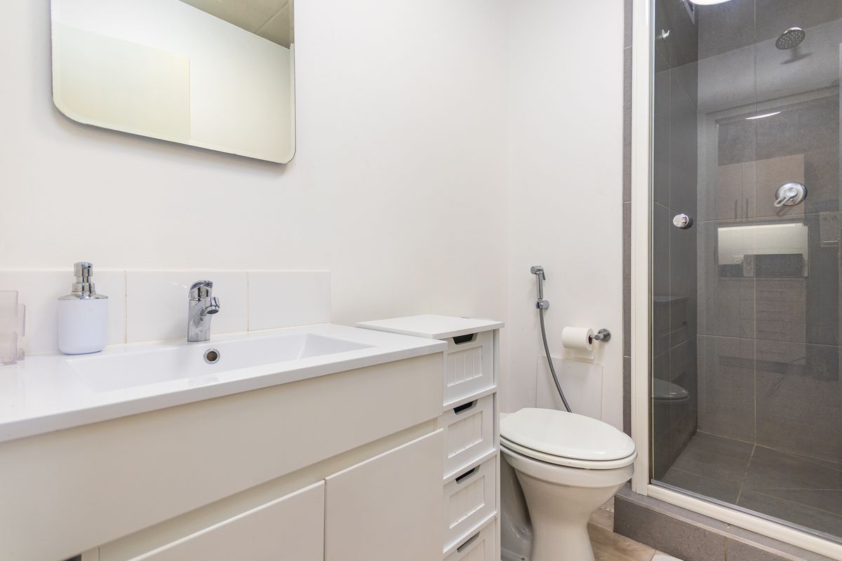 A clean, modern bathroom featuring a white vanity with basin, a wall-mounted mirror, a toilet with bidet spray, and a glass-enclosed shower.