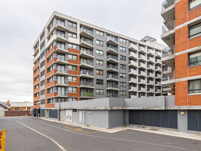 Exterior view of a multi-story apartment complex featuring a mix of brick and concrete finishes with private balconies and ground-level parking access.