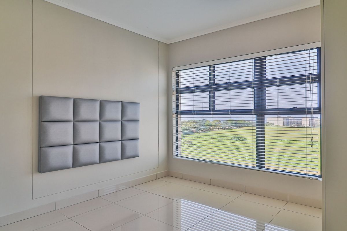 A bright, empty bedroom featuring large windows with venetian blinds, tiled flooring, and a modern upholstered wall panel.