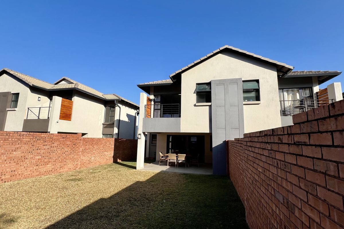 Rear view of a modern two-story house featuring a private garden, brick boundary walls, a covered patio, and an upper-level balcony.
