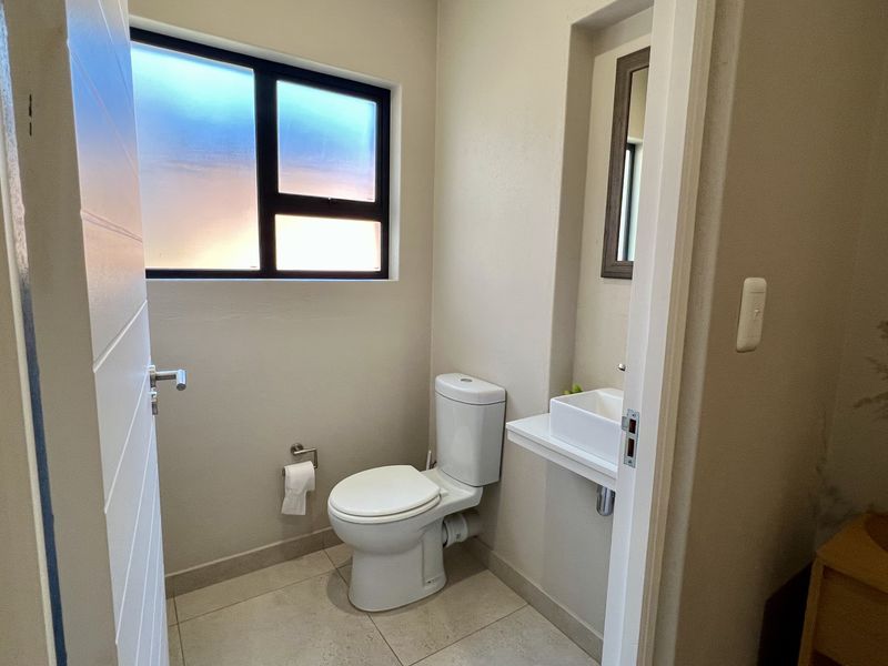 A clean and functional guest bathroom featuring a modern toilet, wall-mounted basin, and neutral tiled flooring with natural light from an obscured window.