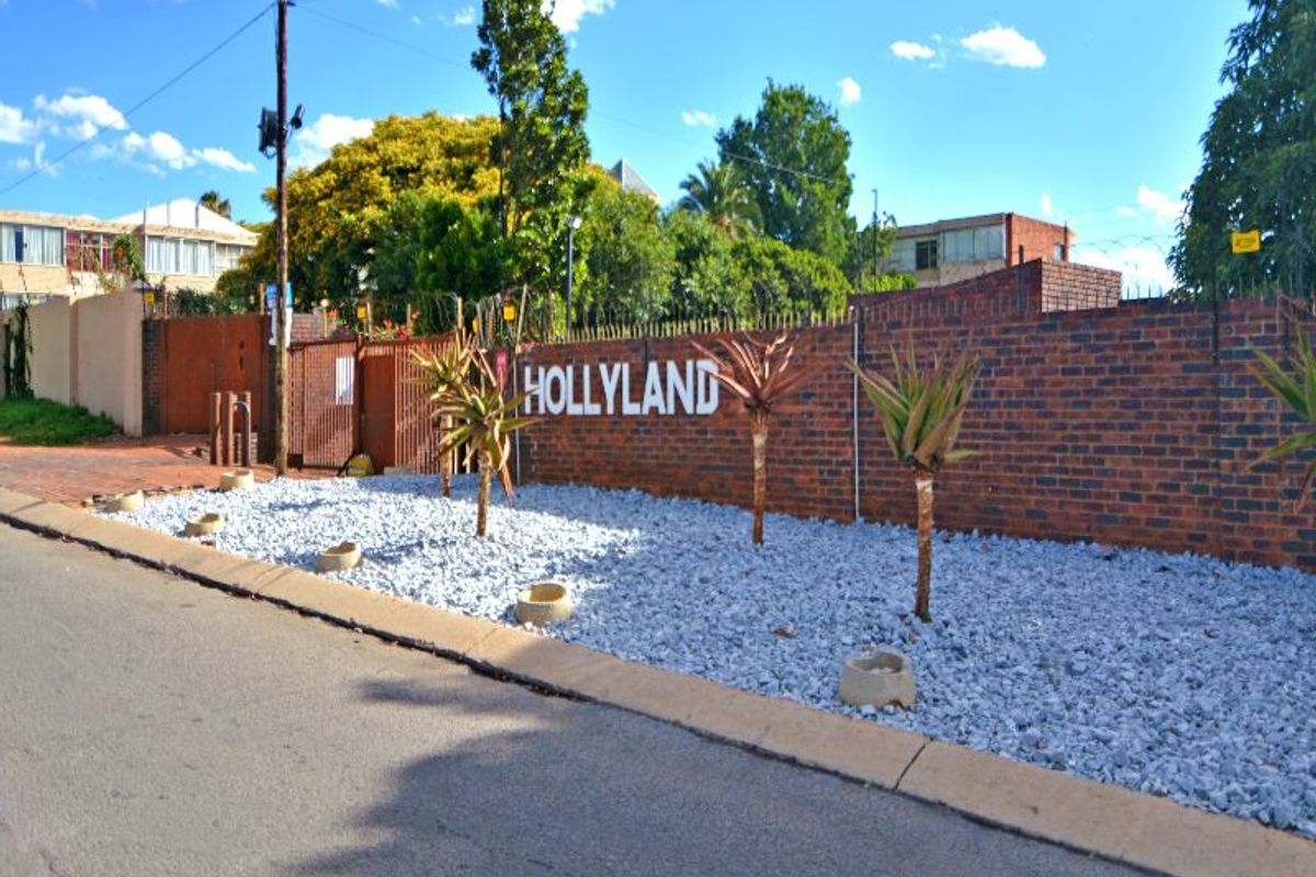 Street view of the Hollyland complex entrance, featuring a brick boundary wall, security fencing, and gated access.