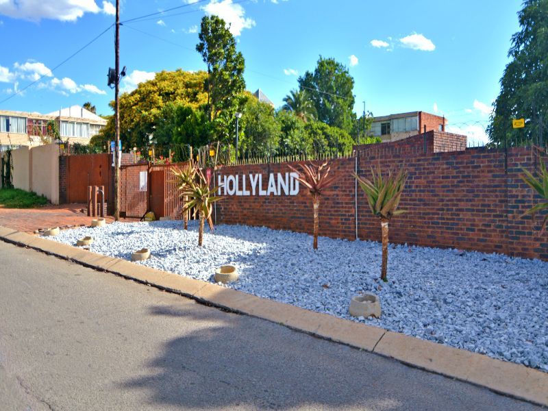Street view of the Hollyland complex entrance, featuring a brick boundary wall, security fencing, and gated access.