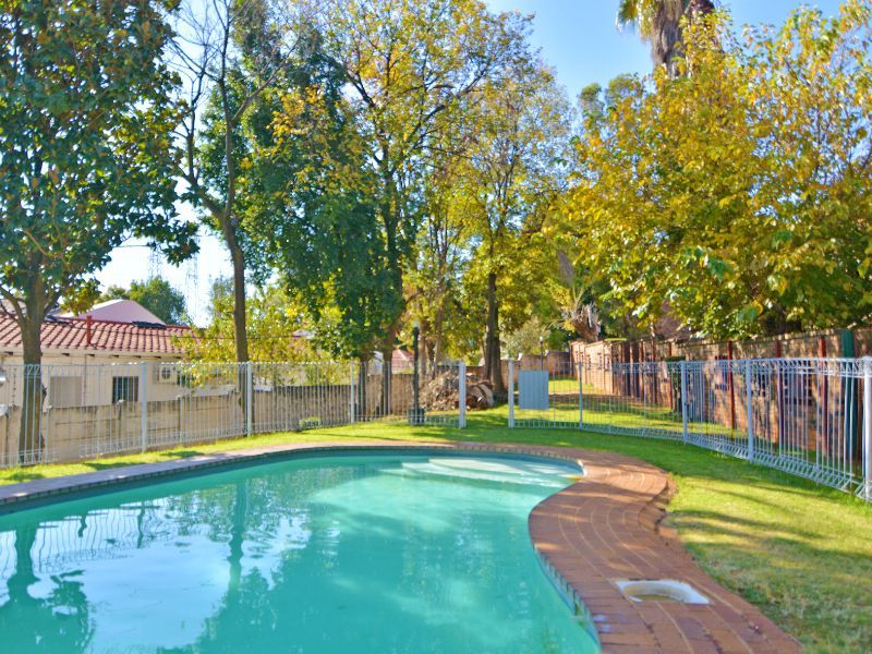 A private swimming pool area featuring brick coping, surrounded by a lawn and metal perimeter fencing within a landscaped garden setting.