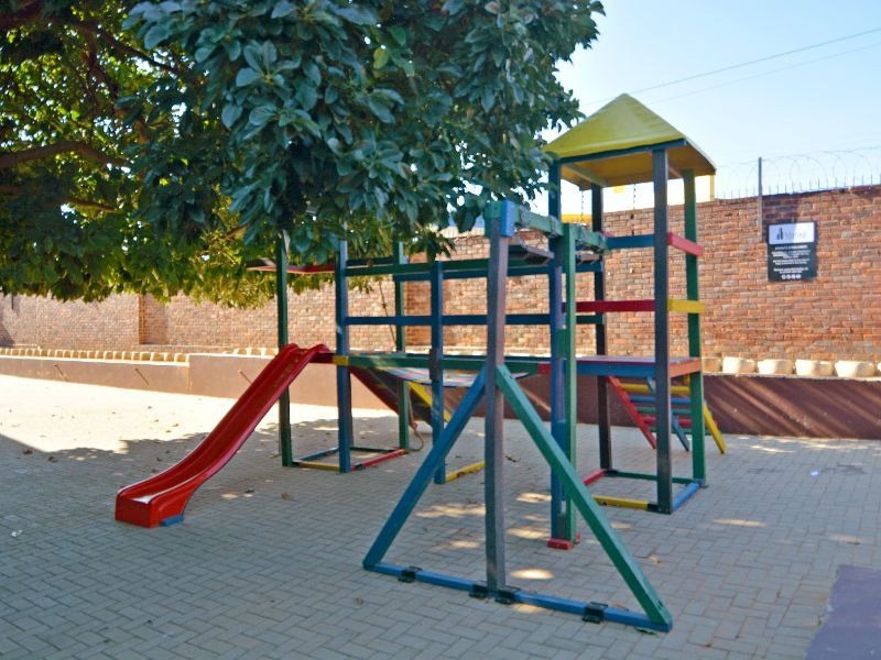 Outdoor communal garden area featuring paved flooring and a fixed children's playground structure, enclosed by a brick boundary wall with security fencing.