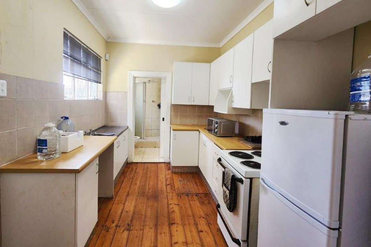 A functional galley-style kitchen featuring timber flooring, laminate countertops, tiled splashbacks, and white cabinetry, with direct access to an adjacent bathroom.