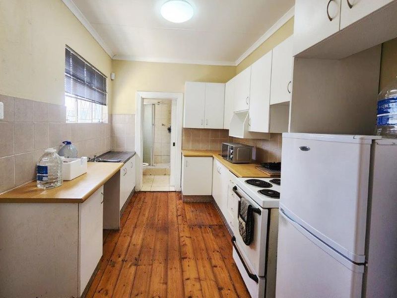 A functional galley-style kitchen featuring timber flooring, laminate countertops, tiled splashbacks, and white cabinetry, with direct access to an adjacent bathroom.