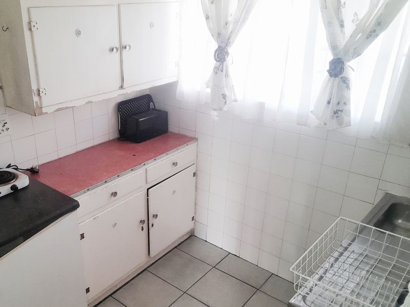 A compact kitchen featuring tiled walls and flooring, with dated built-in cabinetry and a stainless steel sink.
