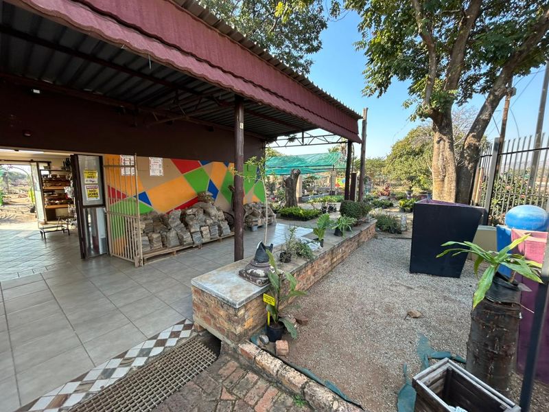 A covered outdoor patio area featuring tiled flooring, a brick retaining wall, and a metal roof structure, suitable for retail or garden display use.
