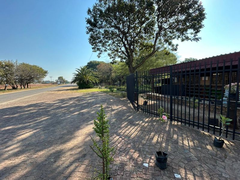 Street-level view of the property entrance featuring a paved driveway, mature trees, and secure metal perimeter fencing.