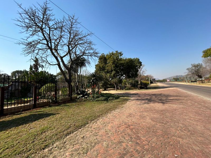 A view of the property frontage featuring a paved driveway, established trees, and secure metal perimeter fencing along the roadside.