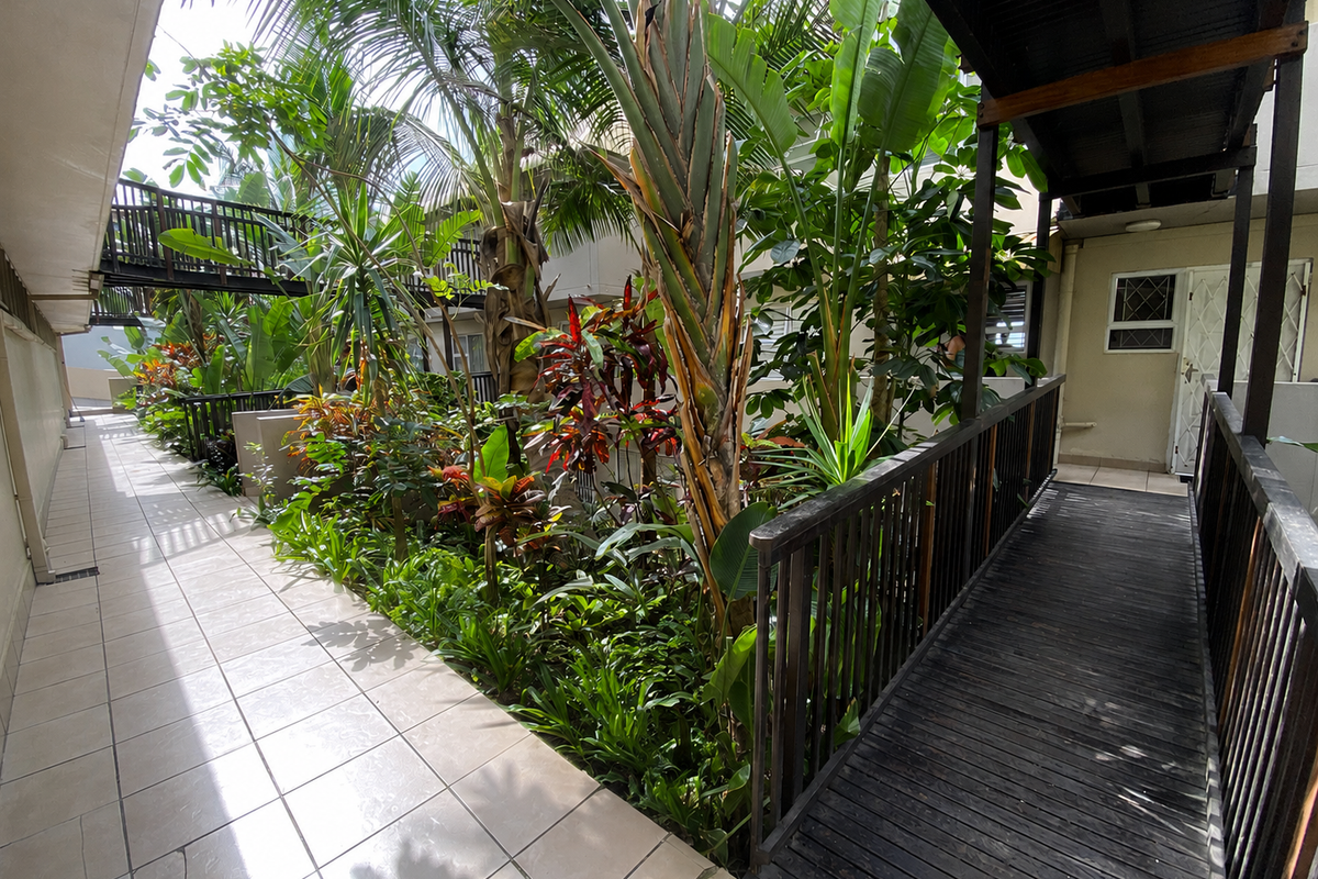 A lushly landscaped courtyard area featuring a tiled walkway, a wooden footbridge, and mature tropical plants between apartment building sections.