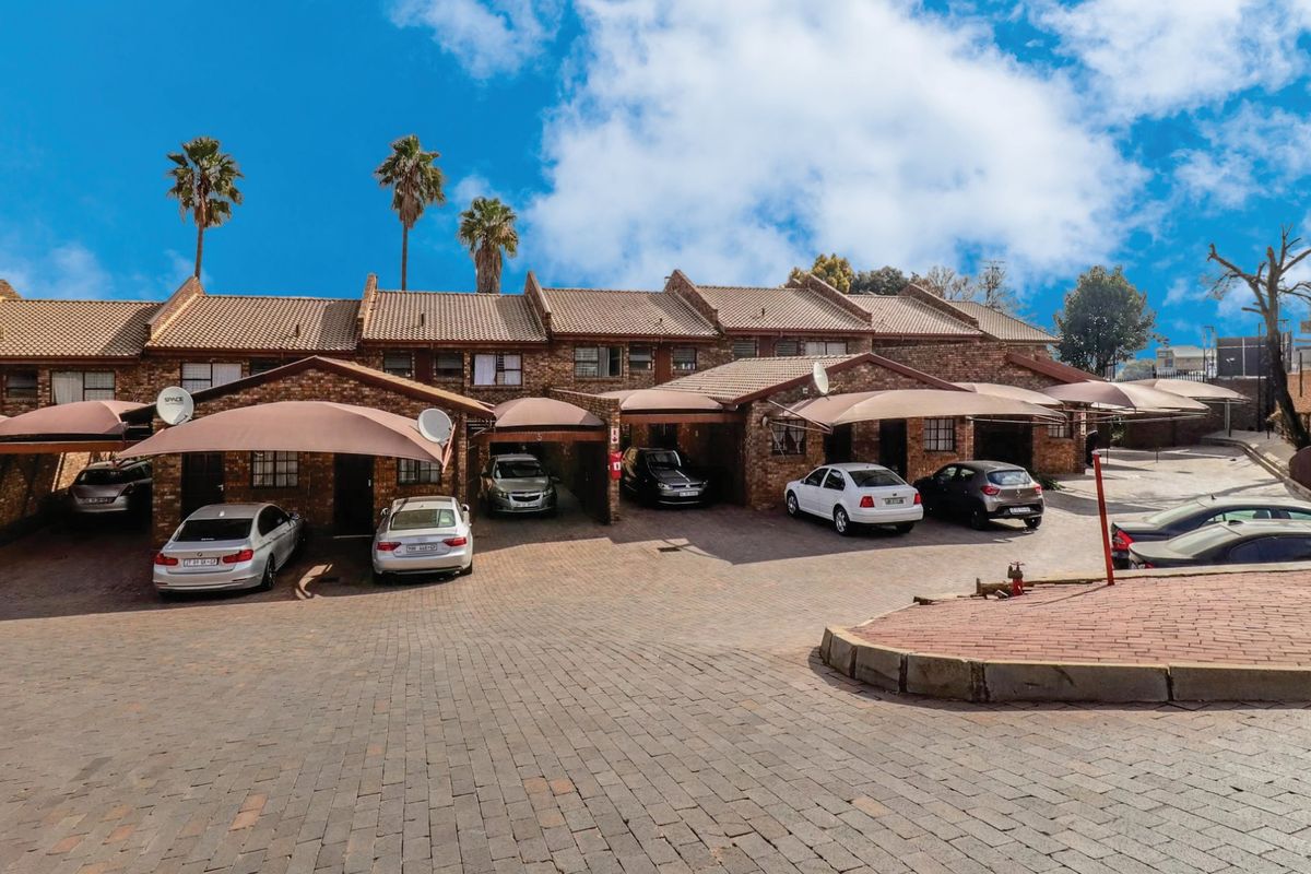 Front exterior view of a townhouse complex featuring brick facades, tiled roofing, and covered parking bays with paved driveway access.