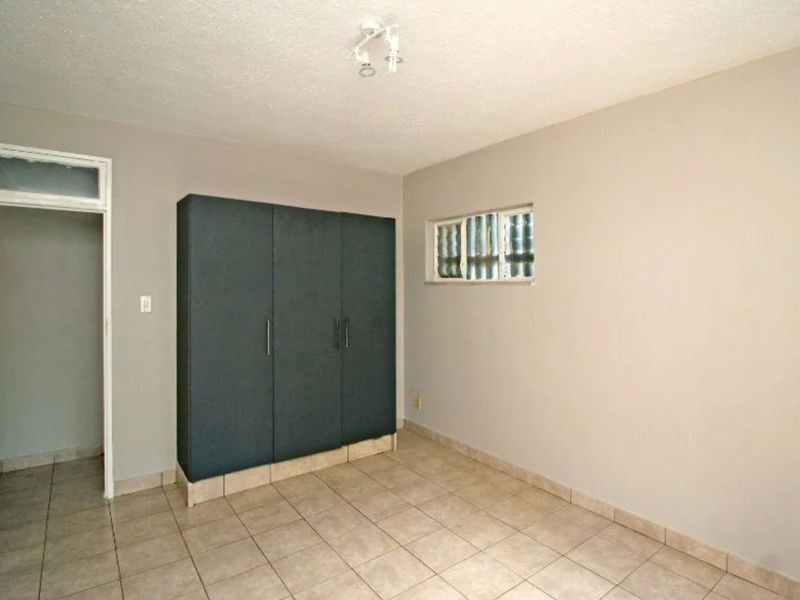 A clean, empty bedroom featuring neutral wall paint, tiled flooring, a built-in wardrobe, and a small glass block window for natural light.